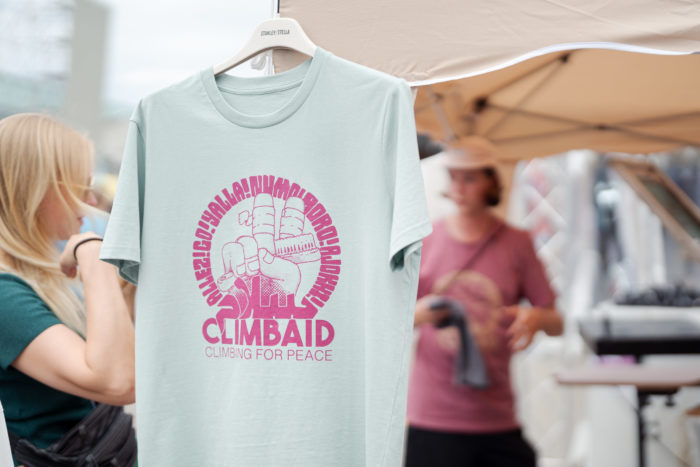 Ein hellblaues T-Shirt, in Pink mit dem ClimbAID-Sujet bedruckt, mit der Hand, die das Peace-Zeichen macht. Zeige- und Mittelfinger sind mit Fingertape umwickelt. Die Hand hält eine Kletterbürste und ist von einem Kreis mit dem Wort für "Los!" in verschiedenen Sprachen umgeben. Im Hintergrund spricht Lino mit einer Kundin.