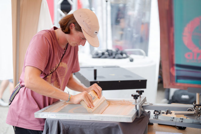 Lino bedruckt ein dunkelgraues T-Shirt, indem er mit dem Rakel die Farbe, ein helles Orange, durch das Sieb streicht. Er arbeitet sorgfältig und konzentriert.