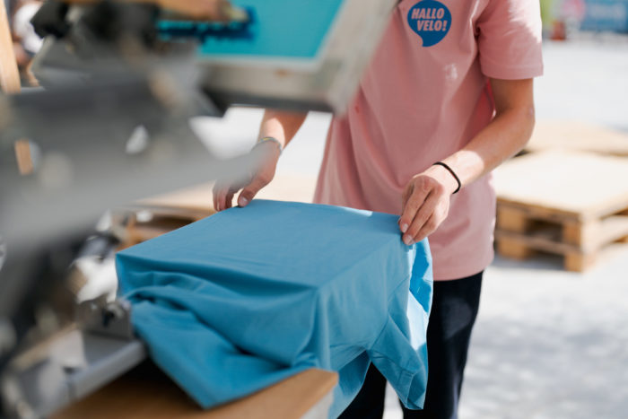 Lino bereitet ein hellblaues T-Shirt fürs Bedrucken vor, indem er es unterhalb des Siebs auf dem Gegenstück glattstreicht. Lino trägt ein lachsfarbenes T-Shirt, das er mit dem Hallo-Velo-Logo in Blau bedruckt hat.